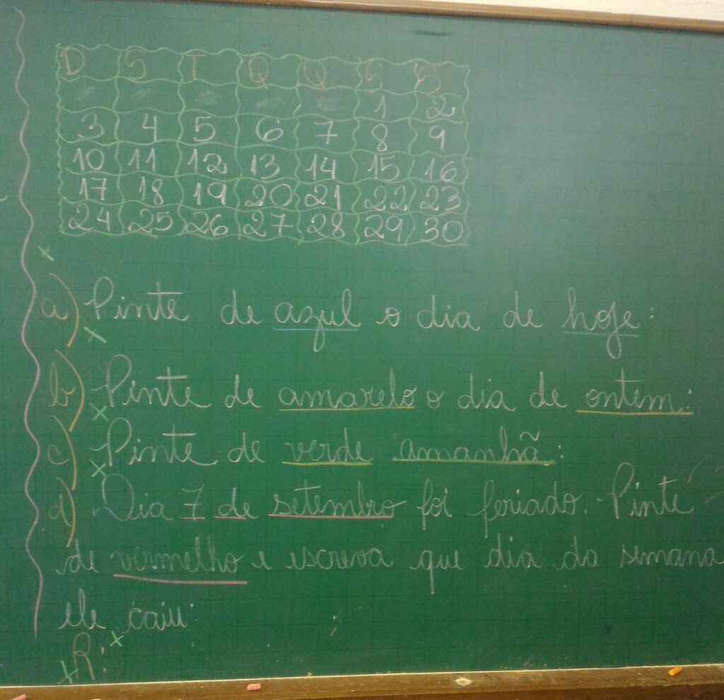 Foto de uma lousa escolar com fundo verde. Nela consta a lição que a professora passou no dia, escrita com letra cursiva. Acima está desenhado o calendário do mês de setembro. Embaixo seguem as questões: A) Pinte de azul o dia de hoje. B) Pinte de amarelo o dia de ontem. C) Pinte de verde amanhã. D) Dia 7 de setembro foi feriado. Pinte de vermelho e escreva que dia da semana ele caiu. Fim da descrição.