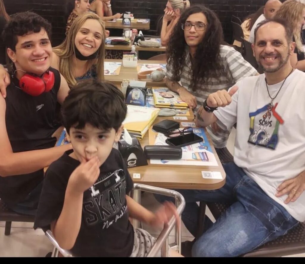 Foto de uma família de 5 pessoas em um restaurante sentados à mesa. O primeiro que está à frente da mesa é o Théo. Ele é um menino de 5 anos, pele branca, cabelo liso, curto, castanho escuro, olhos castanhos, usa uma camiseta preta e está apertando suas bochechas com a mão direita. Atrás dele e à esquerda está seu irmão Rafael, um jovem de 15 anos, pele branca, cabelos castanho-escuros ondulados, olhos castanhos, usa uma camiseta regata preta e está sorrindo. Atrás está a mãe Liliane, mulher de pele branca, cabelos lisos, compridos e loiros, usa uma blusinha de estampas em azul, está sorrindo e com uma máscara preta abaixo do queixo. Do lado direito da mesa, ao fundo, está Lucas, um jovem de 20 anos, pele branca, cabelos compridos e encaracolados, usa óculos, uma camiseta listrada em cinza e branco, esboça um leve sorriso. À sua frente está o pai o Marcelo, um homem de pele branca, calvo na frente e poucos cabelos castanho claro atrás, usa barba curta, uma camiseta branca, está sorrindo e fazendo o sinal de joia com a mão direita. Fim da descrição.